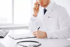 Manlig läkare sitter framför datorn och ringer någon på sin mobiltelefon. Bild från MostPhotos 