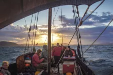 Foto av fören på en båt ute till havs som seglar mot solnedgången. Besättning och tampar i förgrunden.