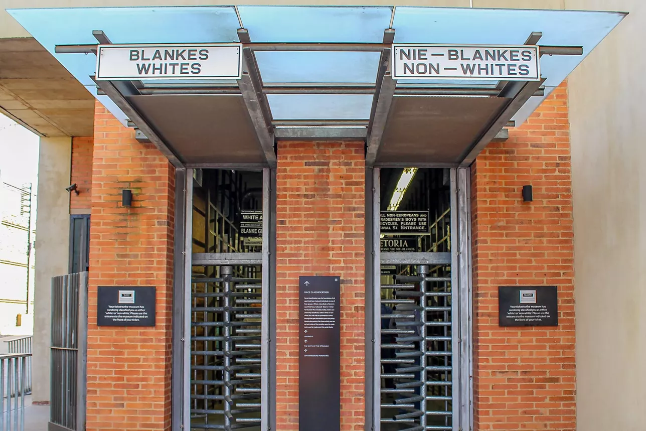 Ingången till en byggnad i tegel. Två smala entréöppningar, en för vita och en för svarta, enligt skyltarna ovanför öppningarna. Byggnaden är Apartheidmuseet i Sydafrika. Foto.