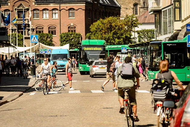 Personer som går och cyklar i Lunds centrum, med bussar och hus runt omkring. Foto.